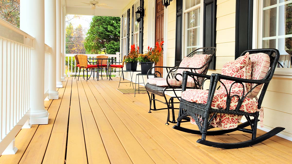 A front porch of a house with white railings and tan floor boards. In the foreground there are two rocking chairs and in the background there is a table with three metal seats surrounding it.